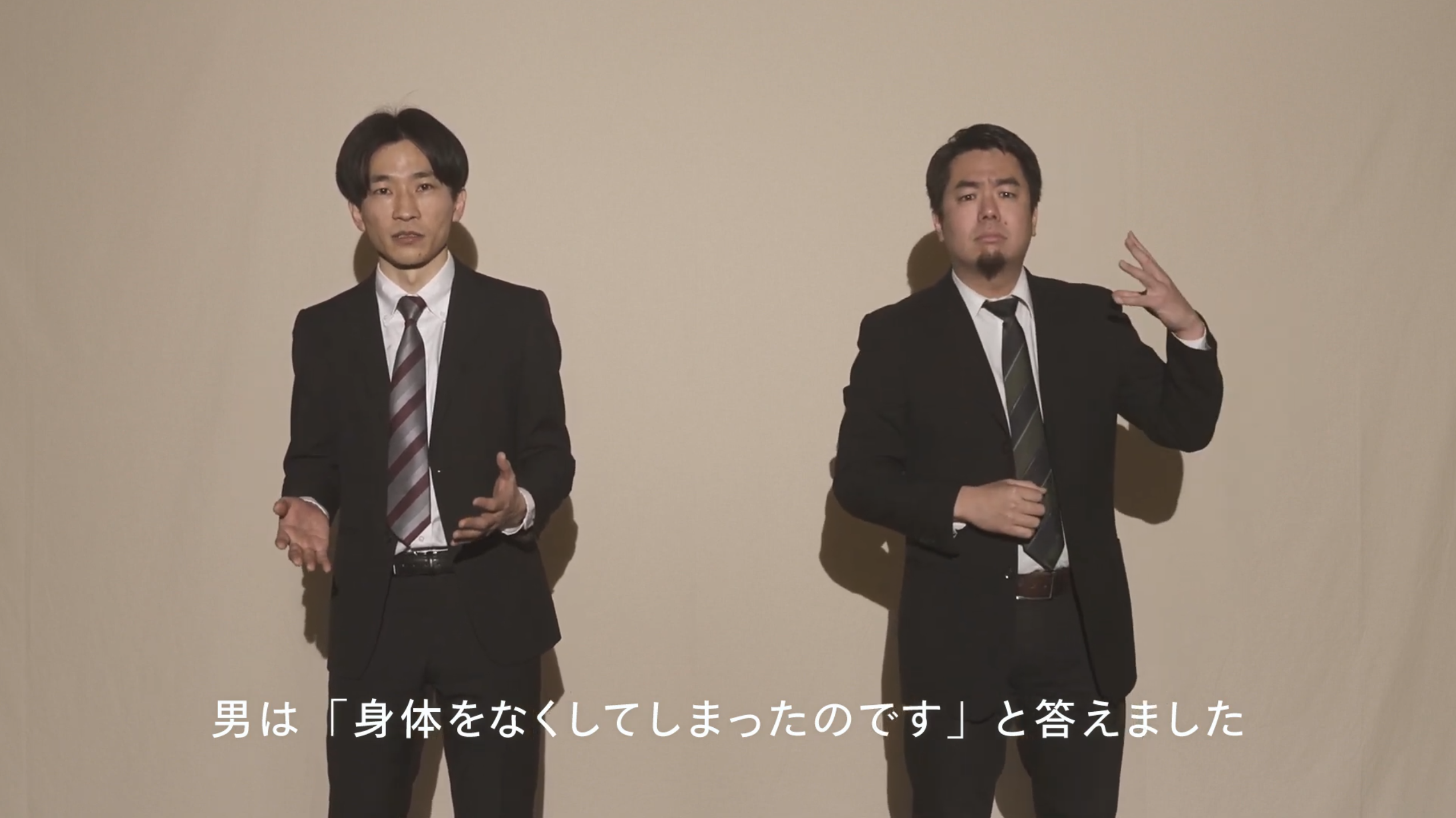 スーツ姿の男性が二人並び、一人は話し、もう一人は手話をしている様子です。画面下部には字幕があります。