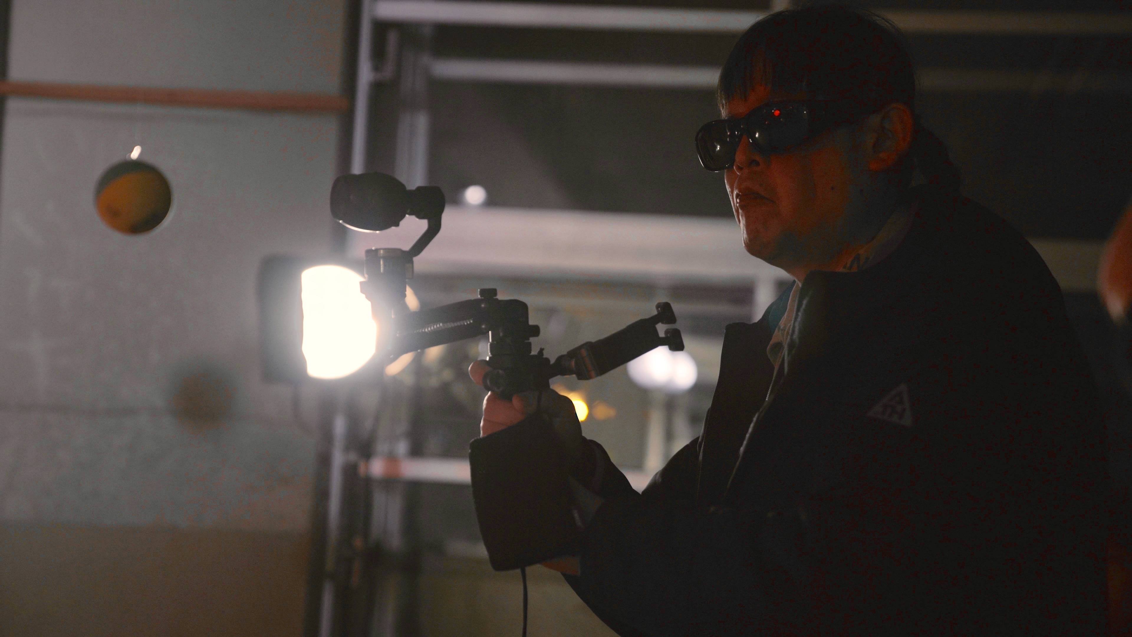 サングラスをかけた男性が、ジンバルに載せたカメラとマイクで、宙に浮いたボールを撮影している。