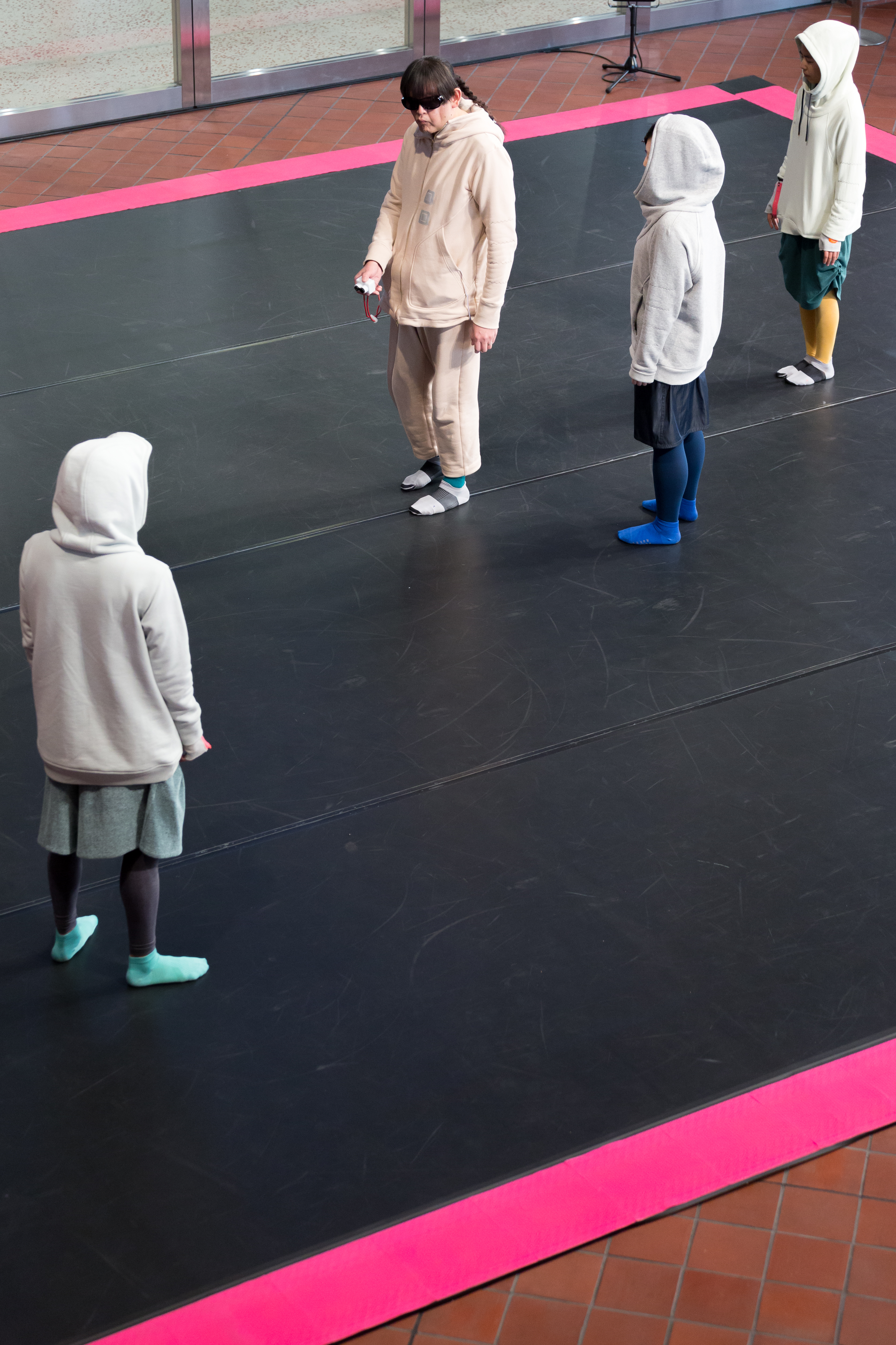 フード付きの4人がピンクの縁取りの黒い床に立っています。1人はアイマスクを着けて、何かを持っています。