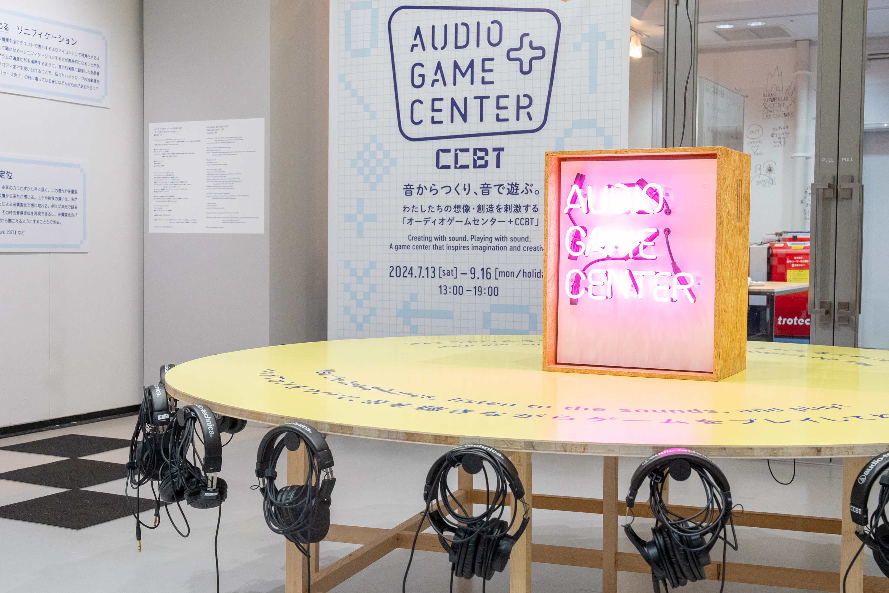 黄色い丸テーブルにヘッドホンが複数ぶら下がり、「AUDIO GAME CENTER」のネオンサインが光る。