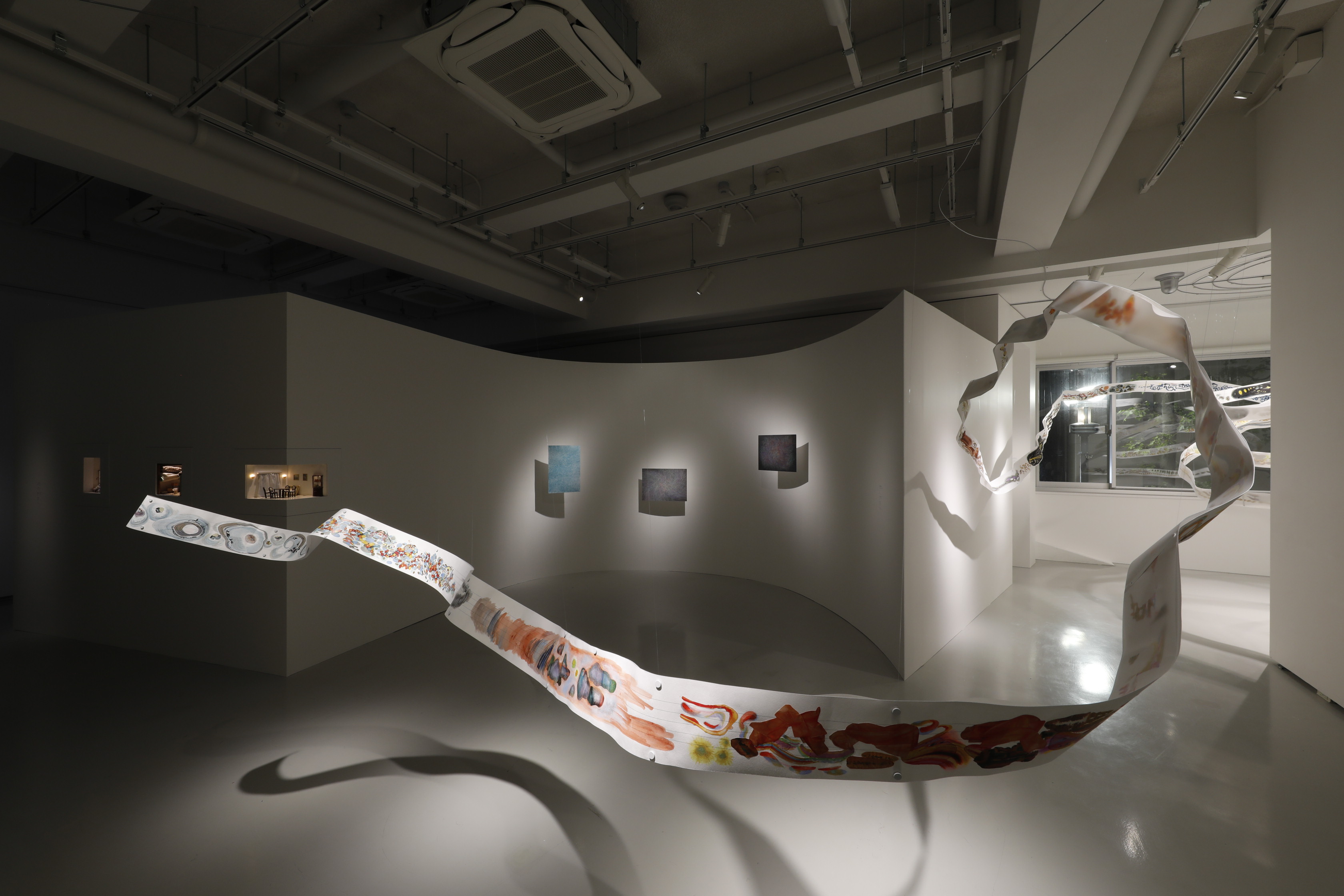 白いギャラリー空間を、抽象画の描かれた長いリボンが空中を蛇行している。壁には絵画数点と、小部屋の模型がある。