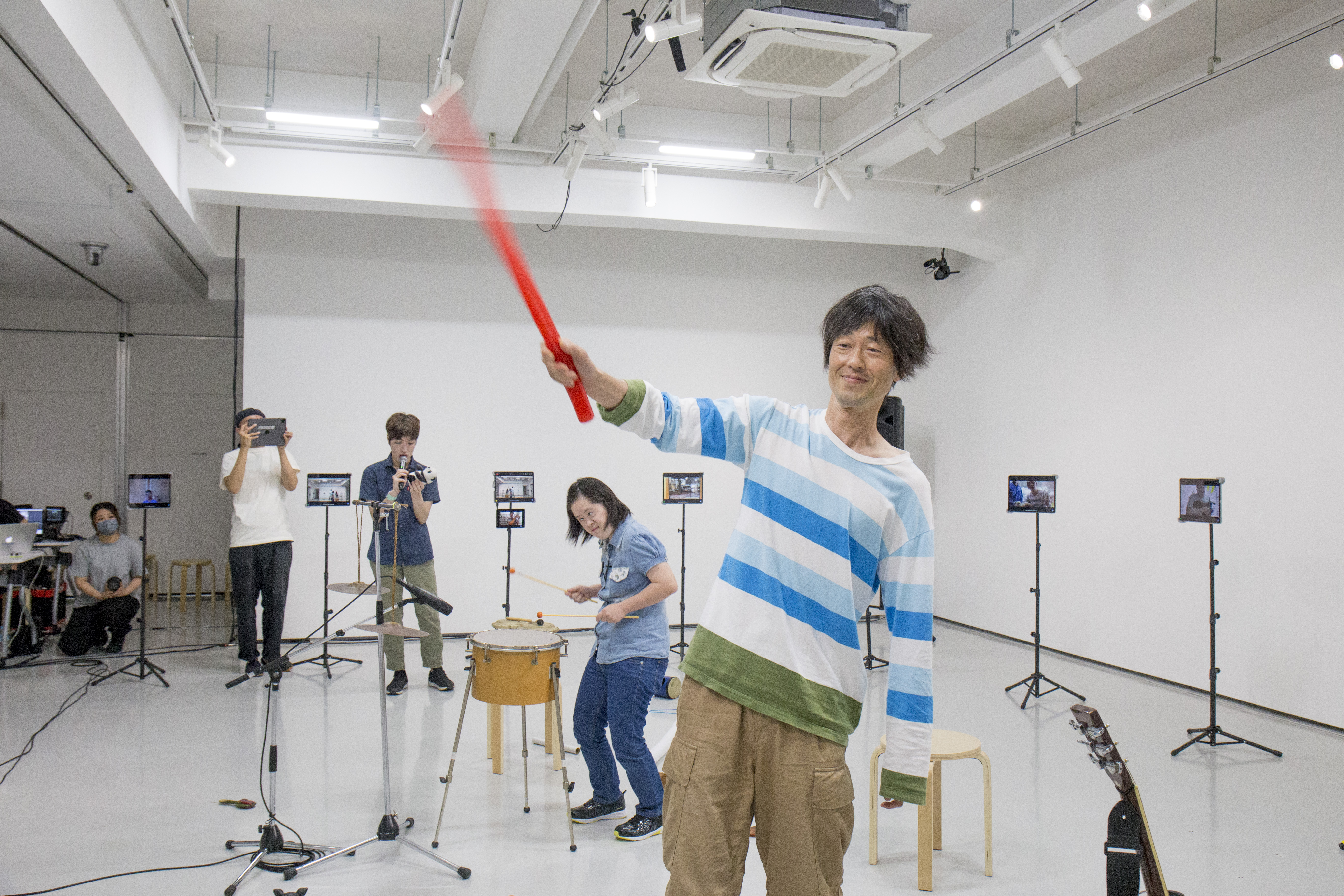 白いスタジオで、ボーダー柄の男性が赤い棒を振り回し、周りに数人が楽器を演奏したり、タブレットで撮影している。