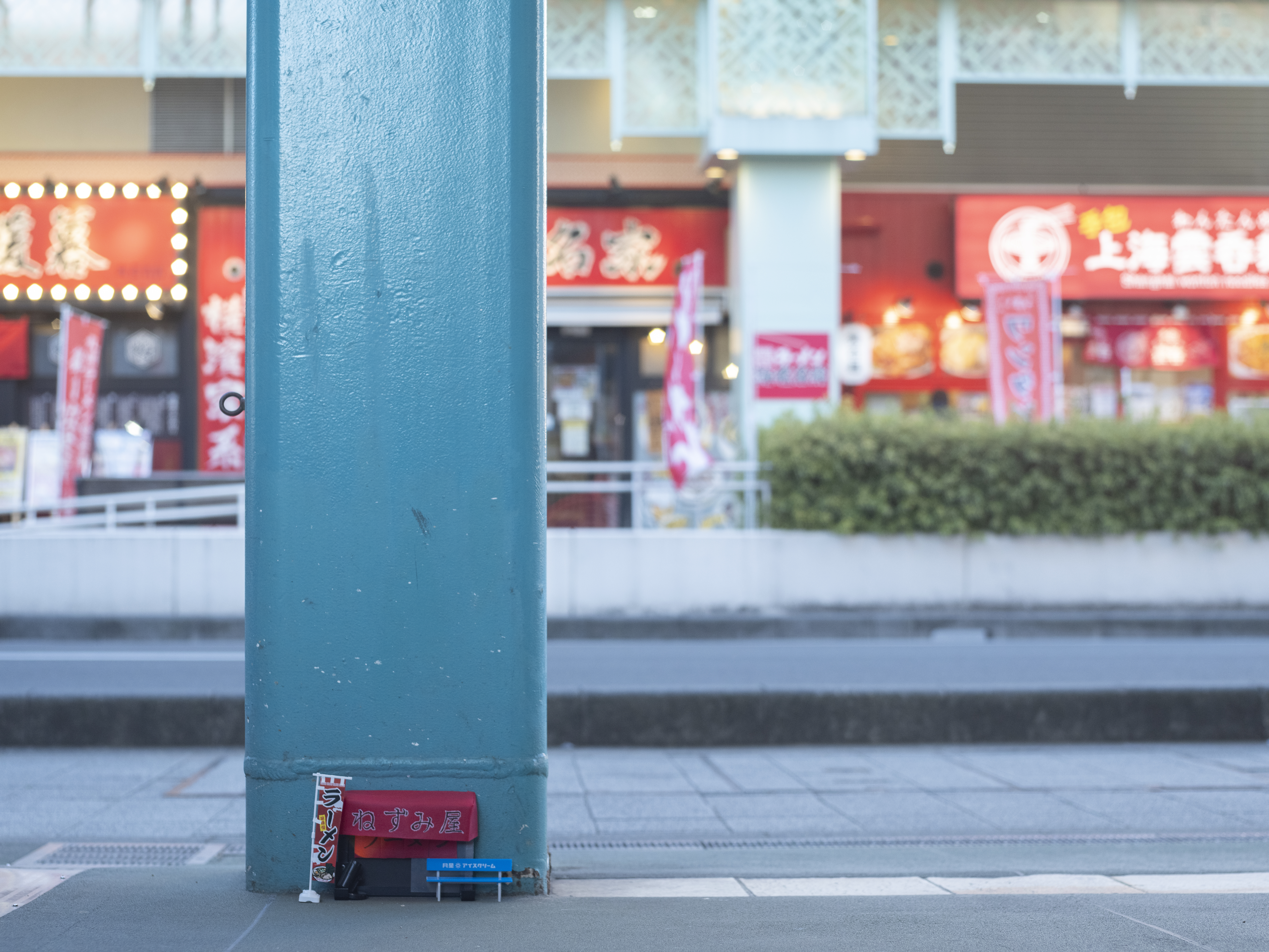 青色の柱の根元に、ミニチュアのラーメン屋台。赤い幟と小さな青いベンチがある。背景にぼやけた飲食店街。