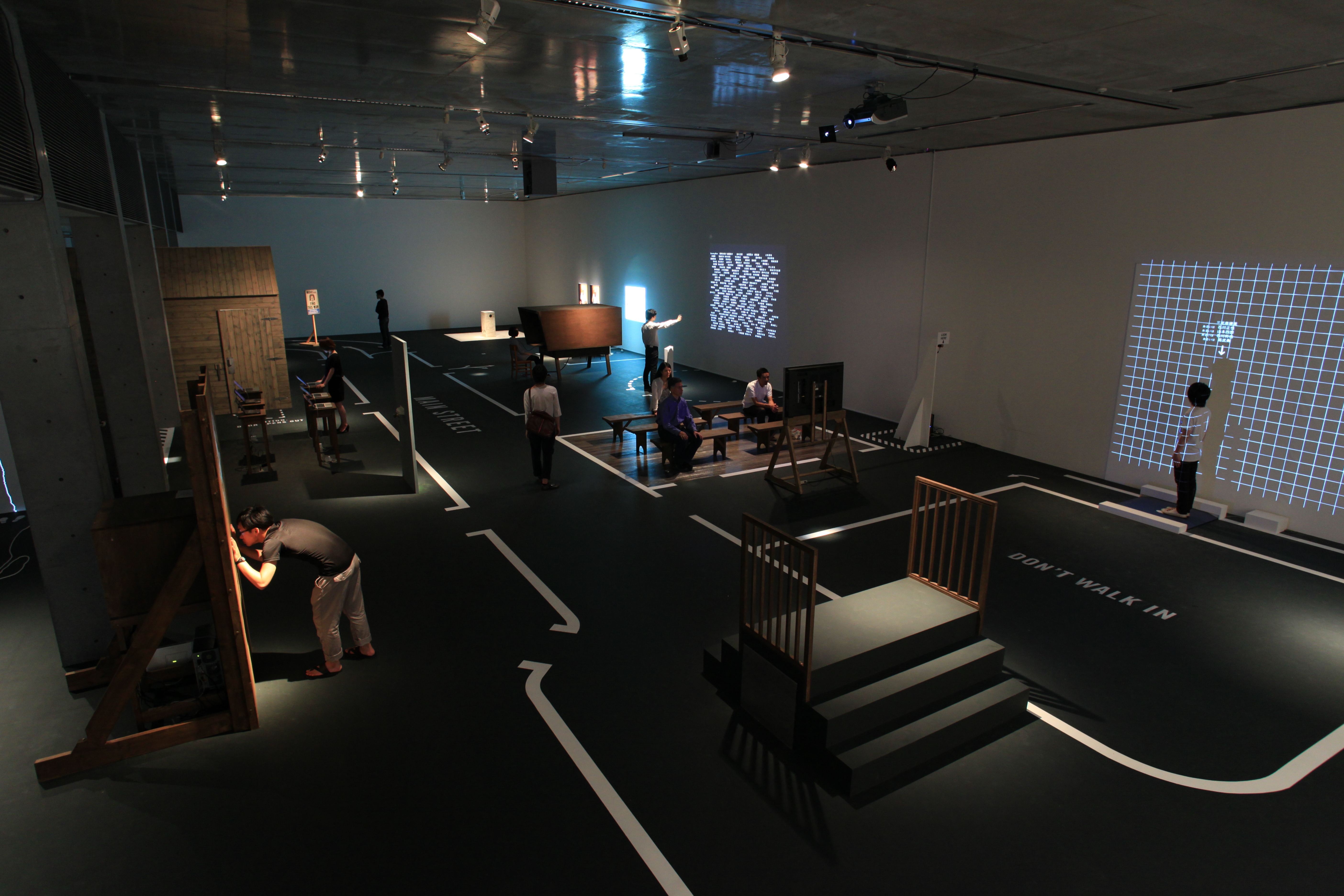 暗い展示室。白い線で区切られた床に、人がインタラクティブな展示物を見たり操作したりしている。プロジェクションマッピングあり。